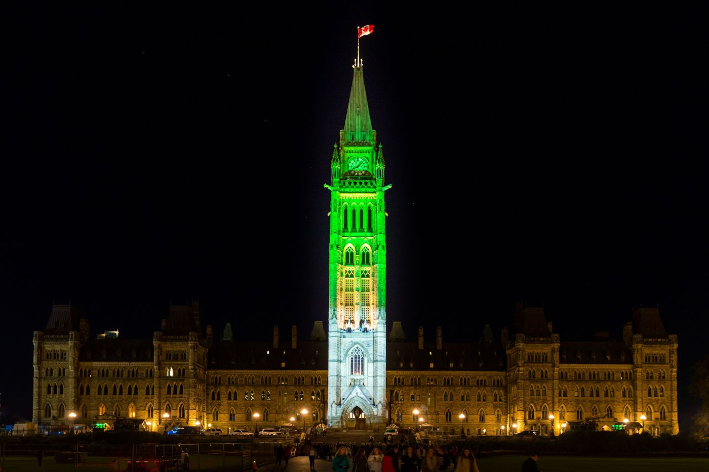 " Colour photograph of a large, six-storey building at night. A Canadian flag flies at the top of the tower in the centre. The tower is illuminated in blue and green."