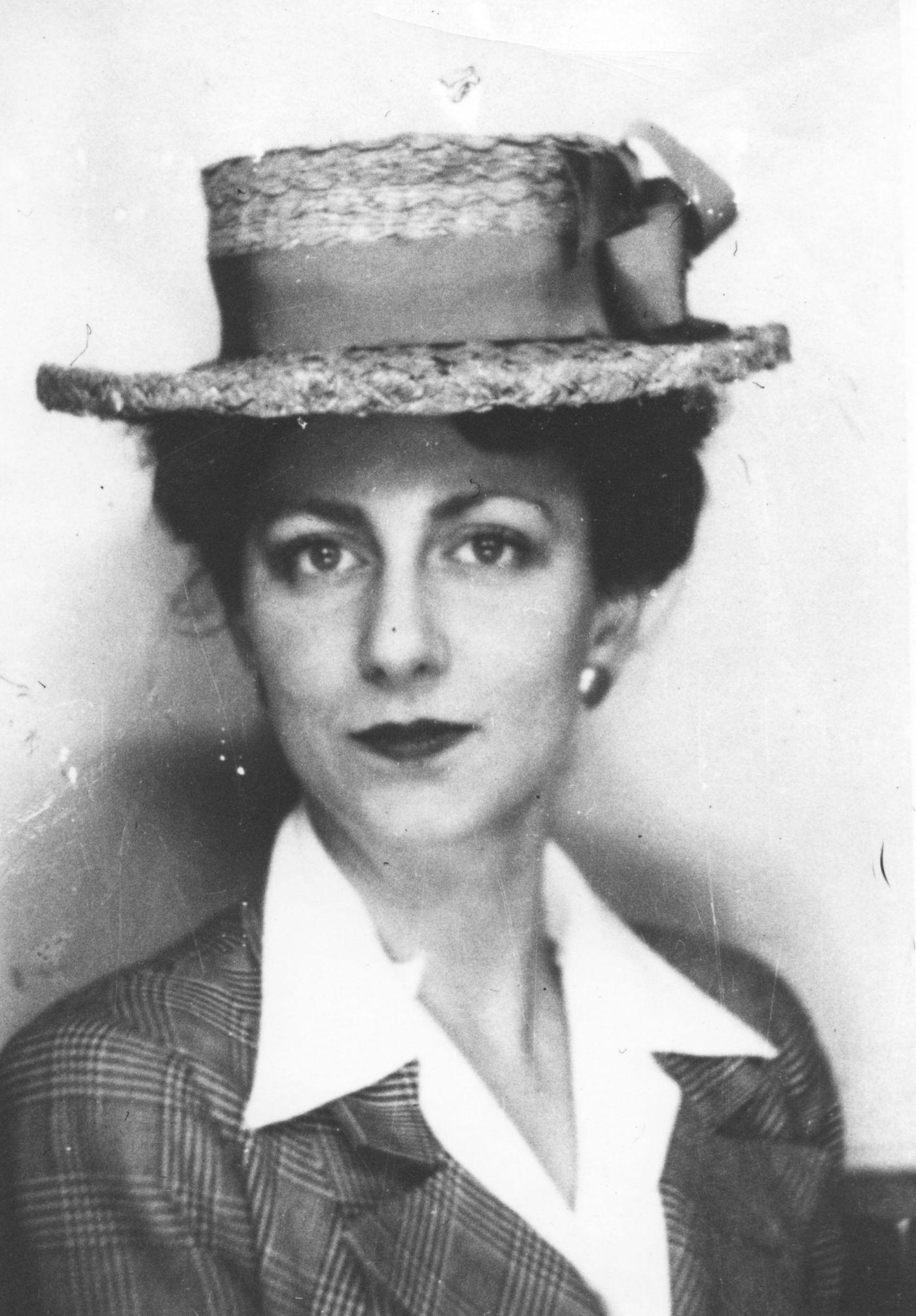 Portrait en noir et blanc d’une jeune femme. Elle a le regard très direct. Elle porte un élégant chapeau de paille décoré d’un large ruban ainsi qu’un veston de plaid et un chemisier blanc.