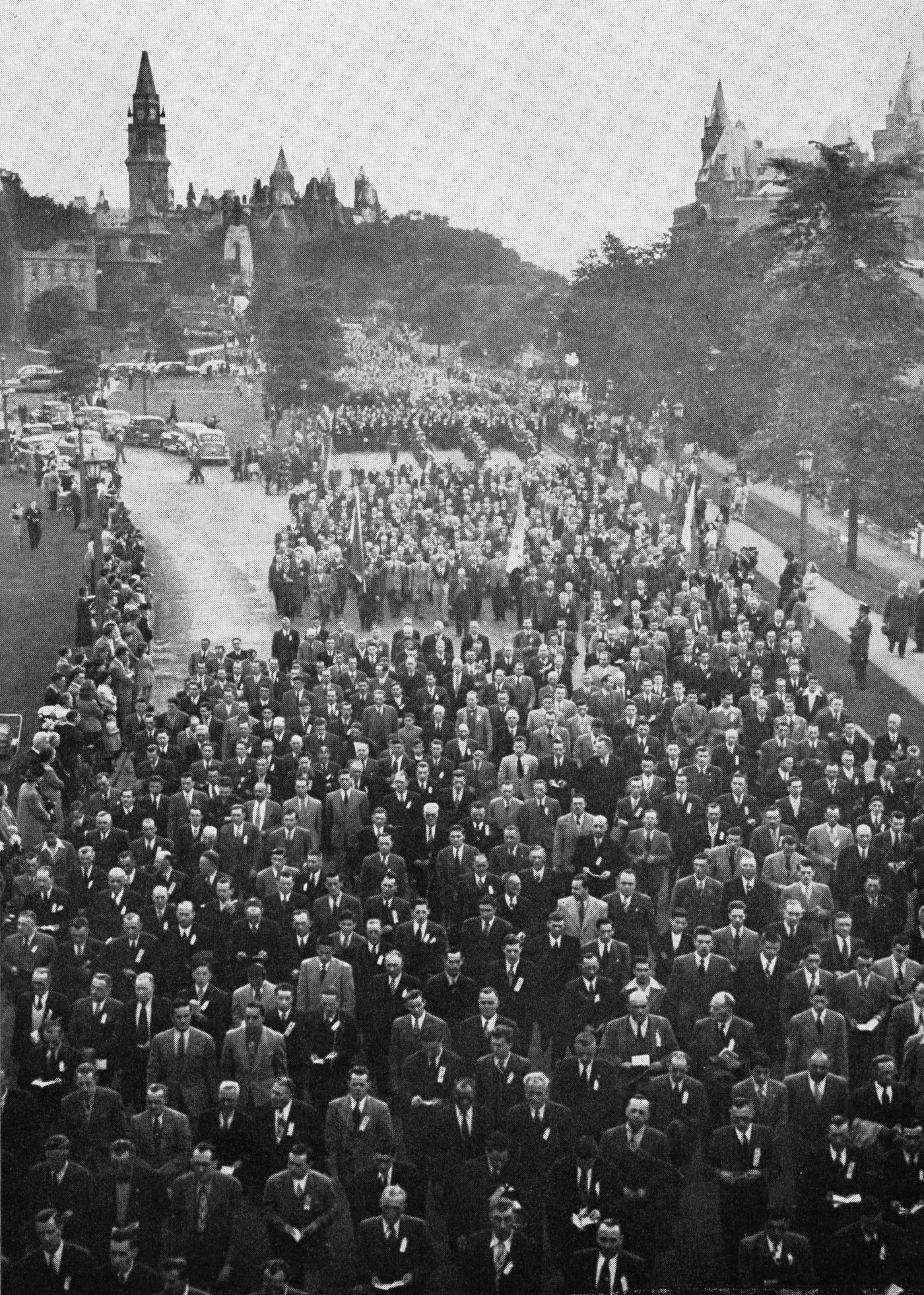 Black and white photograph of a grand procession. Men in suits and ties are walking down a roadway, some holding booklets, others holding flags. Spectators are massed along the route. In the distance  behind them, buildings of Parliament.