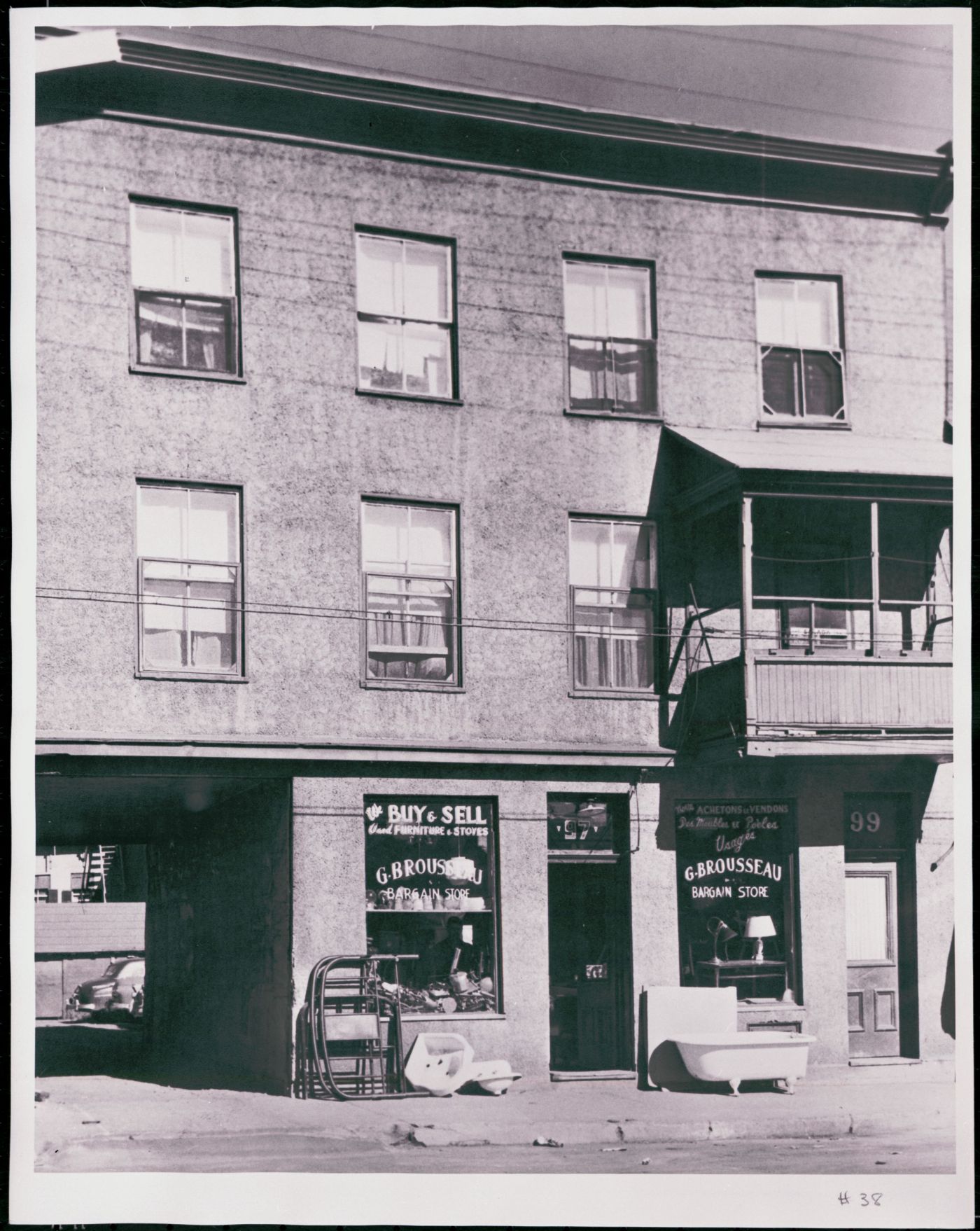Photographie en noir et blanc d’un édifice à trois étages en stuc, avec un balcon au premier étage. Au rez-de-chaussée, un magasin d’objets usagés avec affichage bilingue à la devanture. Un passage permet d’accéder au stationnement à l’arrière de l’édifice.