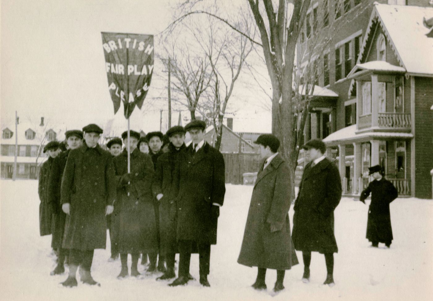 A small group of people dressed in winter clothing stand outside, in the snow in an open space surrounded by brick buildings and trees. One of the group members holds a flag with English text reading “BRITISH FAIR PLAY PLEASE“ in capital letters.