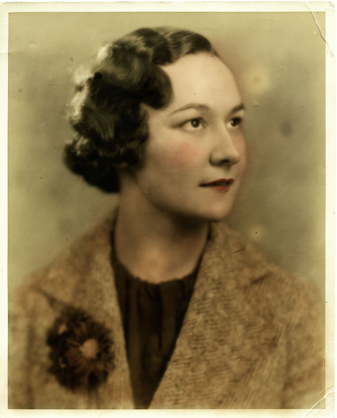 Colour studio photograph showing the head and shoulders of a middle-aged woman in three-quarter profile view. She has short, wavyhair and wears a jacket with flowers in the buttonhole.