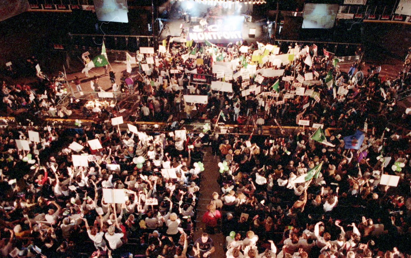 Photographie en couleur d’une foule importante, au pied d’une scène. Il est écrit « S.O.S. Montfort » sur le plancher de la scène. La foule brandit des drapeaux franco-ontariens ainsi que des ballons verts et blancs et des pancartes affichant le slogan « S.O.S. Montfort ».