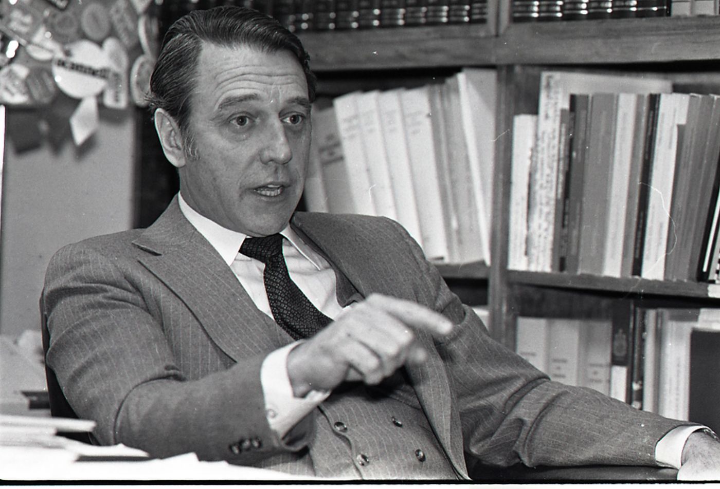 Black and white photograph, three-quarter view, of a dark-haired, middle-aged man wearing a grey pin-striped suit, white shirt and dark tie. He is speaking, pointing with his index finger. Behind him, a bookshelf and part of a bulletin board.