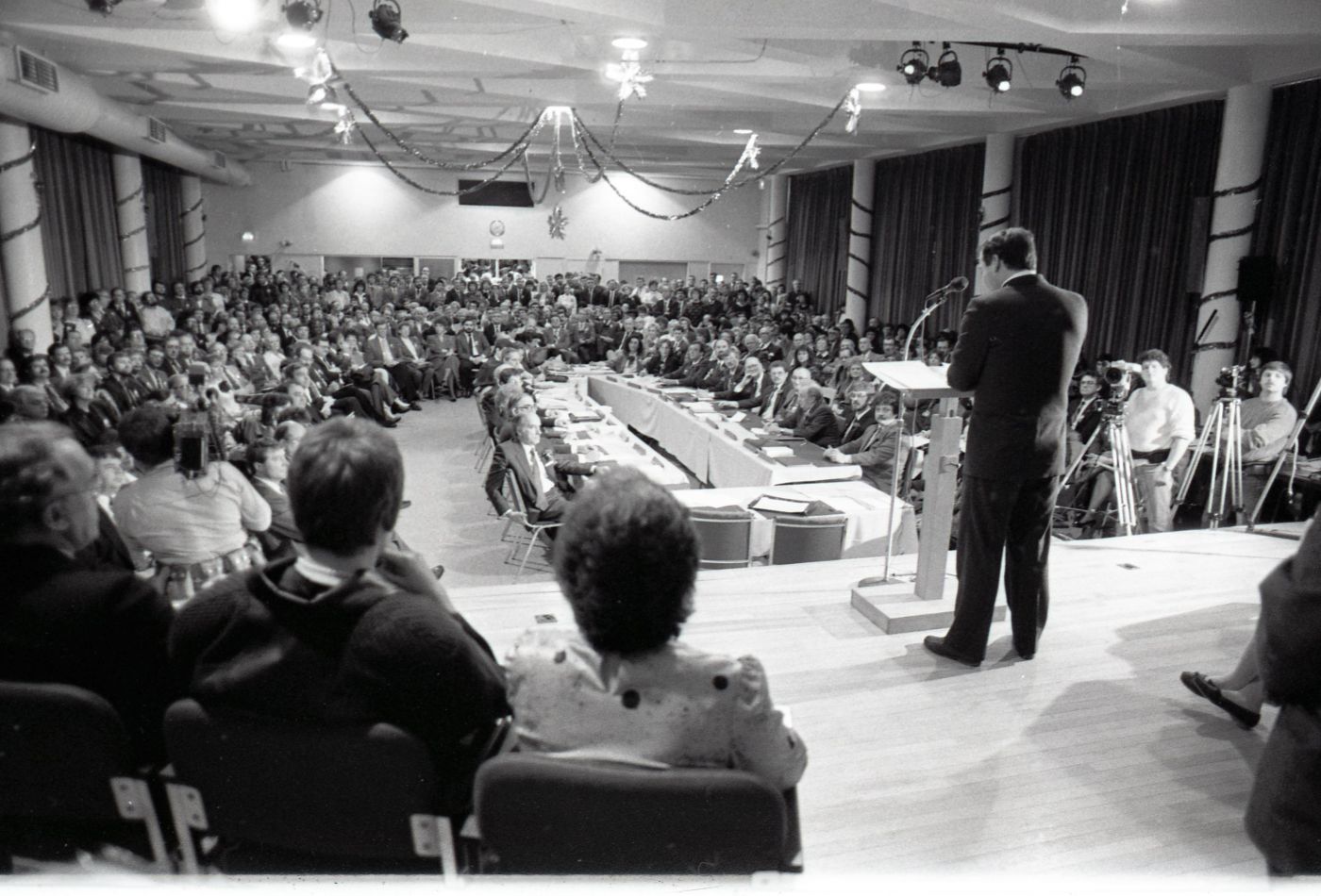 Photographie en noir et blanc d’un homme vu de dos en costume, debout sur une scène et parlant au microphone. Il s’adresse à un public nombreux dans une salle comble et à des journalistes.