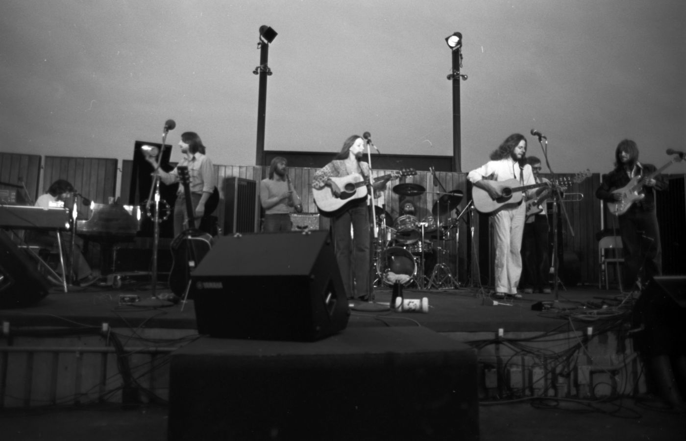 "Black and white photograph of seven musicians (three guitarists, a bassist, a pianist, and two percussionists) on an outdoor stage in dim light. The group includes six long-haired men and a woman, of mature age, wearing casual clothes.  "