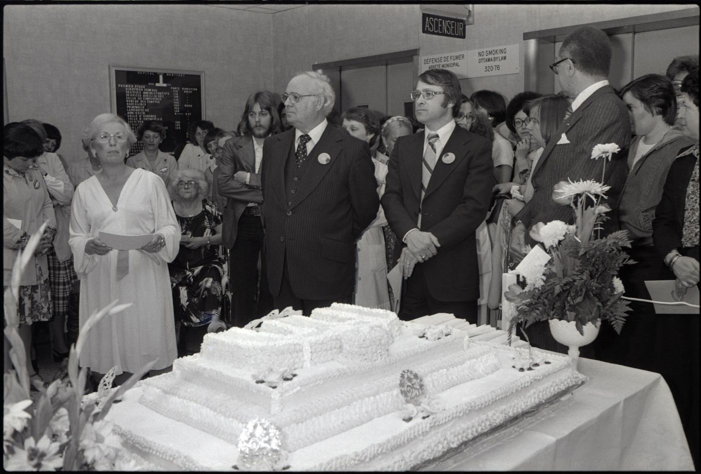 Black and white photograph of a group of people standing around a huge cake decorated with the logo of the Montfort Hospital. Two wreaths of flowers are placed on the table. In the foreground, three men in suits wearing the same button, and an elegantly dressed woman holding a sheaf of papers.