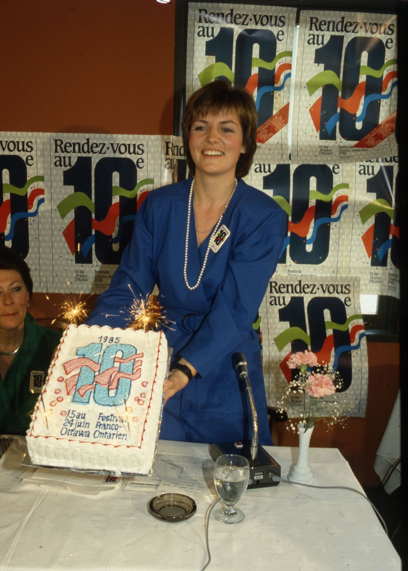 Colour photograph of a middle-aged, smiling woman with short brown hair, wearing a blue dress. She holds up a cake decorated with a large number 10, a date, and the words “Festival Franco-Ontarien.” Behind her, a wall covered with 10th anniversary promotional posters.