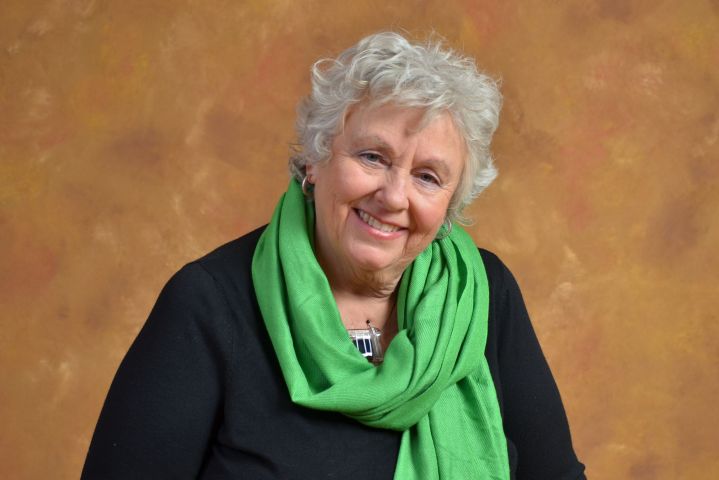 Colour studio photograph of an older woman with curly gray hair. She wears a black jacket, with a green scarf tied around her neck. She is smiling.