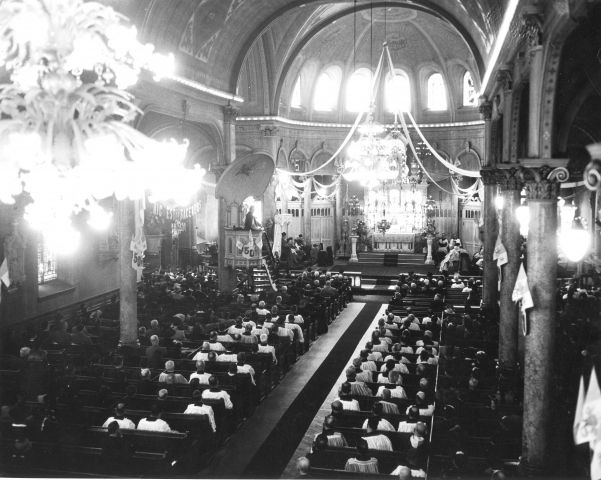 Black and white photograph of the interior of a church seen from above. The church is richly decorated. Attendance is strong.