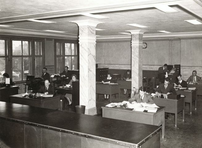 Black and white photograph of the interior of an open concept office. Seen from the front of the room, eight men and four women sit at desks, working. Large windows line one of the walls.