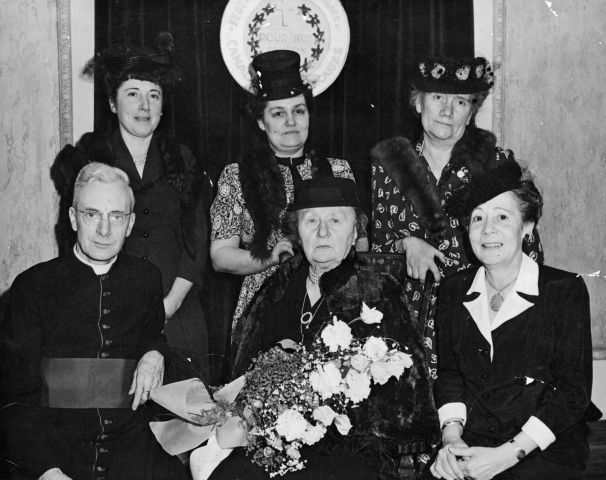 Black and white photograph of a group of women of various ages, wearing hats and dresses and fur stoles, except for one. Two women are seated, and one holds a bouquet of flowers. A priest is seated to their right. Three other women are standing behind them.