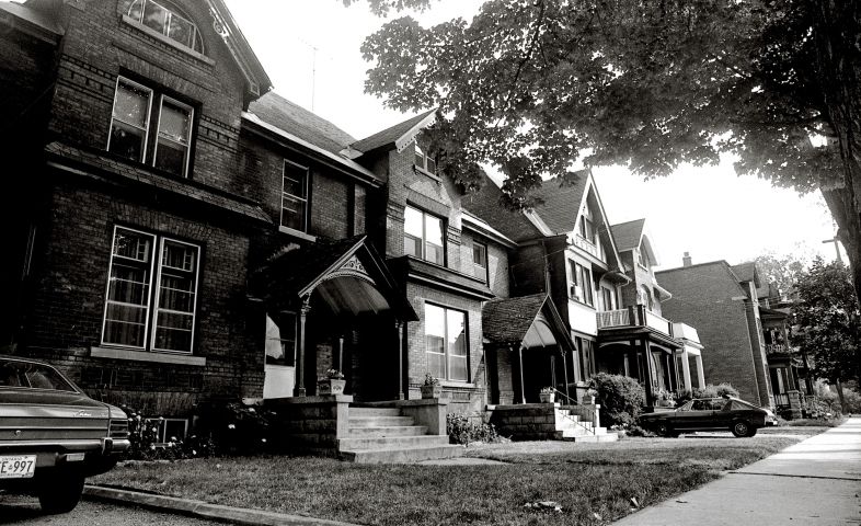 Black and white photograph of several adjoining three-story brick houses with basement, sidewalk views. Cars are parked in two of the driveways. The street is filled with trees, and flowers grow in the small, well-maintained yards.