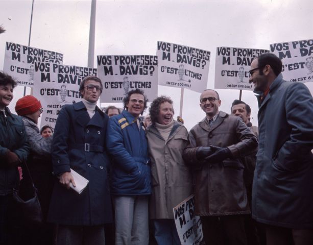 Colour photograph of five middle-aged people – four men and a woman – in front of a group of demonstrators. They smile and hold placards which read, “Vos promesses, M. Davis ? C’est l’temps. On est 600 000 !” (Your promises, Mr. Davis? It’s time. There are 600,000 of us!).