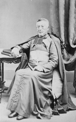 Black and white photograph of an elderly man wearing the liturgical garments of a bishop. He does not wear the cap. He sits on a chair, his right arm resting on a table, his hand resting on books.