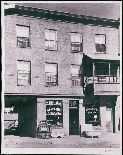 Black and white photograph of a three-storey stucco building with a balcony on the first floor. On the ground floor, a used goods store with bilingual signage at the front. A passageway offers access to the parking lot at the rear of the building.