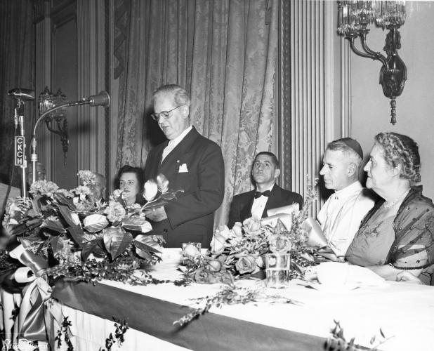 Black and white photograph of a mature man in a suit and tie, standing and talking into a microphone at a decorated table. A woman is seated to his right. Sitting to his left: a man, a cleric wearing a skullcap, and another woman.