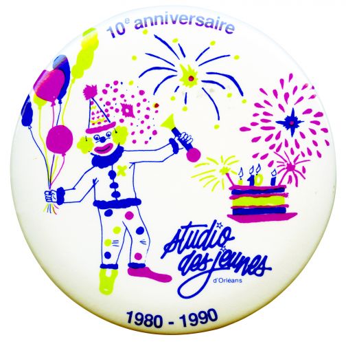 Photograph of a button featuring on white a clown holding balloons, a birthday cake and fireworks in bleu, light green and fuhsia. A blue logo appears on the button as well as an acknowledgement of the anniversary.