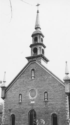 Black and white photograph of a brick church, surmounted by a bell tower and two turrets. The facade of the church has three doors, a bull’s-eye window, and three statues.
