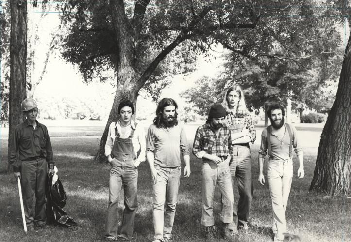 Black and white photograph of a group of five young men in a wooded park. Three have beards, and two have long hair. They wear casual clothing – suspenders, overalls, chequered shirts – and they are walking toward the camera. Next to them, an older man picks up garbage.