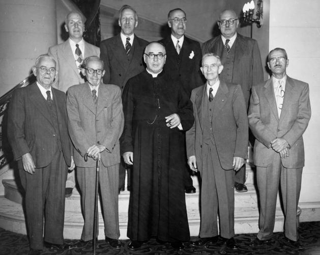 Black and white photograph of nine elderly men. Five men stand at the bottom of a staircase, and four stand behind them on the second step. Eight of the men are dressed in suits and ties. The man in the centre is dressed in a black cassock and holds a cigar. One of the men in the front row leans on a cane.